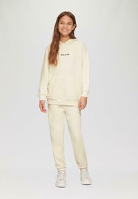 Helles Creme-Zweiteiler-Outfit, bestehend aus einem Kapuzenpullover mit einer Vorderentasche und passenden Jogginghosen, beide aus weichem, strukturiertem Stoff gefertigt.
