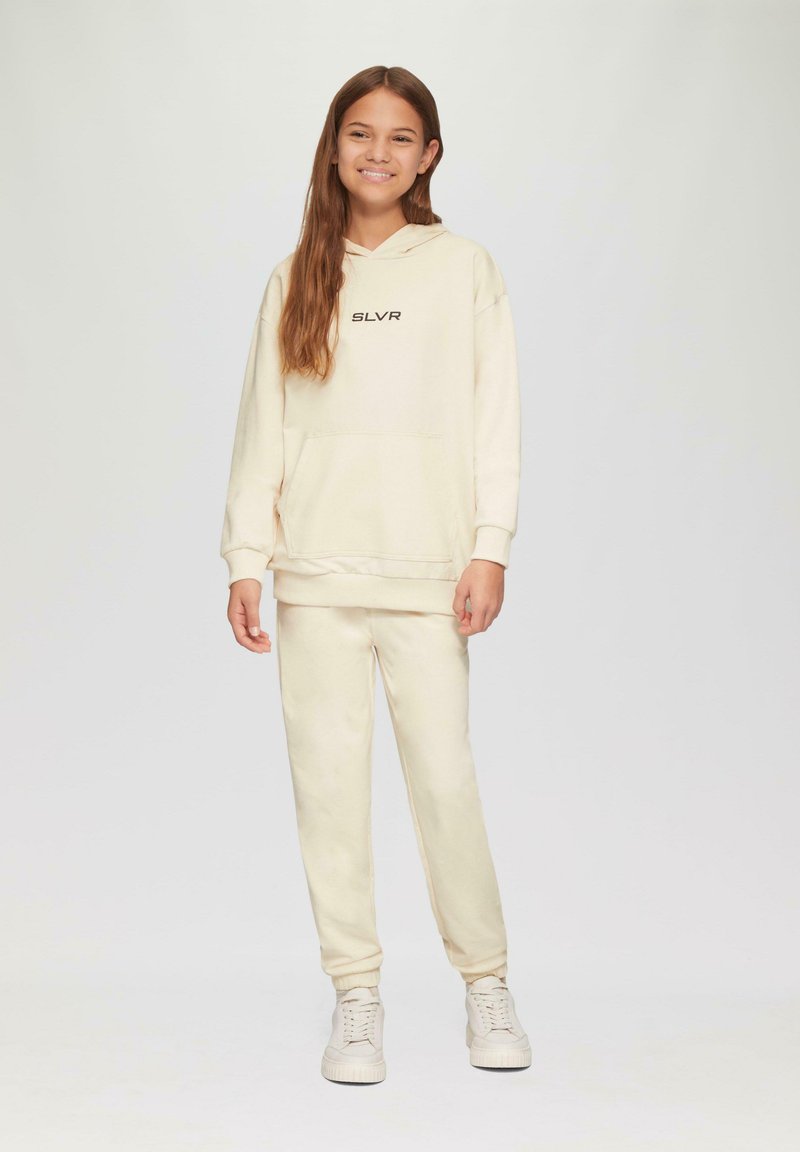 Helles Creme-Zweiteiler-Outfit, bestehend aus einem Kapuzenpullover mit einer Vorderentasche und passenden Jogginghosen, beide aus weichem, strukturiertem Stoff gefertigt.