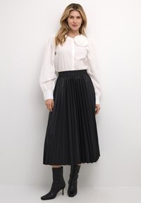Biela blúzka s button-up uzáverom, s nafúknutými rukávmi a kvetinovým detailom; čierna plisovaná midi sukňa; čierne vysoké členkové čižmy.