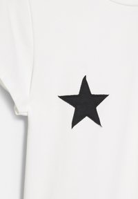Witte t-shirt van katoen met een zwart, stervormig patch aan de linkerkant, met een gladde textuur en korte mouwen.