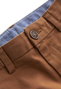 Gros plan sur la ceinture d'un pantalon marron avec le bouton fermé, montrant les passants de ceinture et le tissu de doublure intérieure bleu.