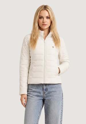 Junge Frau mit langen blonden Haaren, die eine weiße gesteppte Jacke mit Reißverschluss und hellblaue Jeans vor einem schlichten Hintergrund trägt.