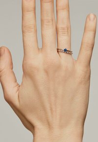 Conjunto de anillo de oro con una piedra preciosa azul en el centro rodeada de pequeñas piedras claras, colocado en una mano de tono de piel claro.