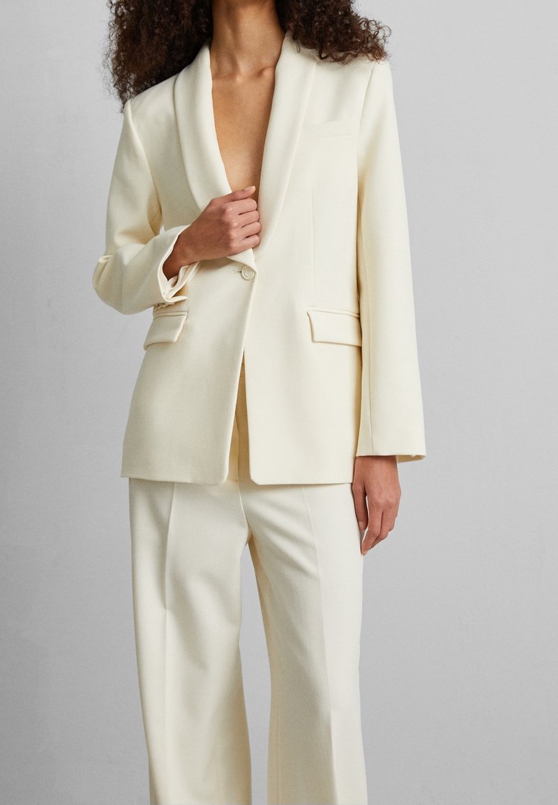 Femme aux cheveux bouclés portant un blazer sur mesure blanc cassé et un pantalon assorti, main ajustant le blazer près de la poitrine.