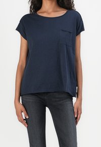 Námořnická modrá krátkosrstá tričko vyrobené z měkkého materiálu, s kapsou na hrudi a volným střihem, spárované s tmavě šedými džíny.