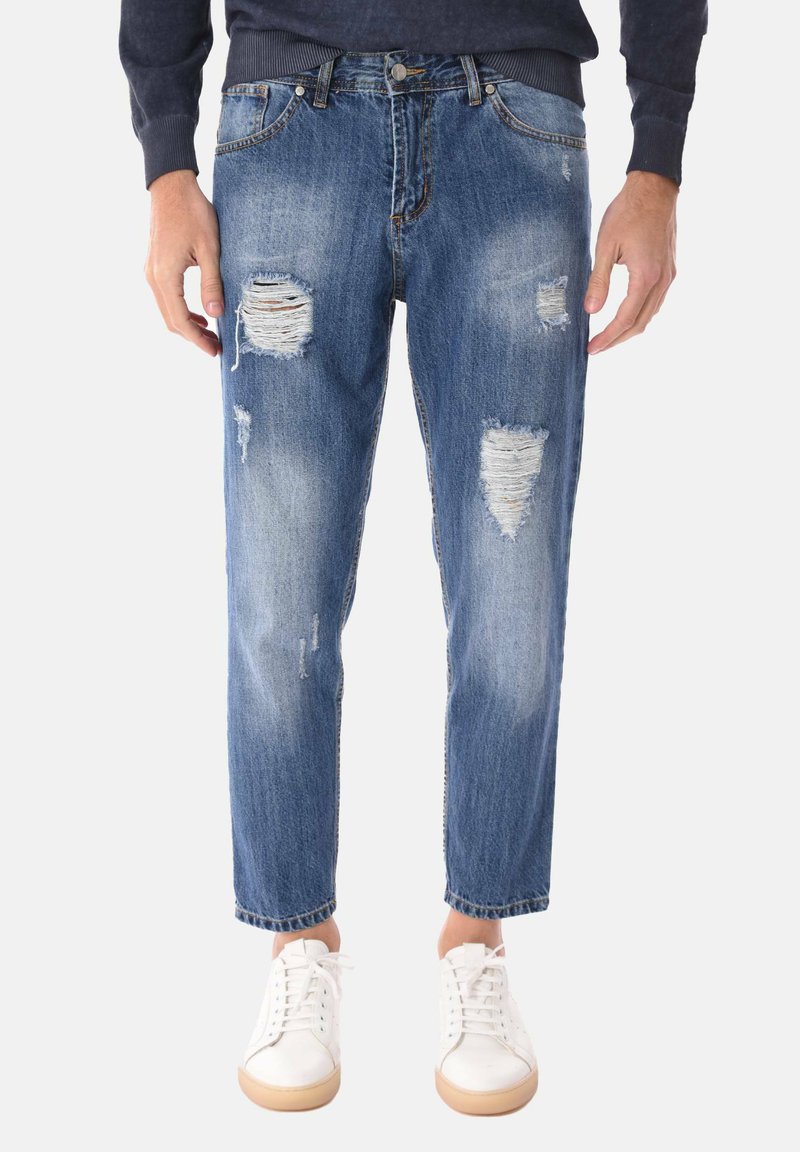 Jeans blu usurati con molteplici strappi indossati con scarpe da ginnastica bianche e un maglione scuro su uno sfondo neutro.