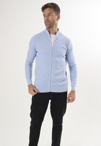 Ljust blå stickad zip-up kofta med ribbad textur, sidofickor och hög krage, kombinerad med svarta byxor. Slät tygfinish.