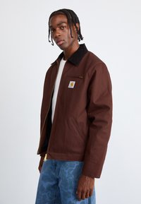 Brown canvas jacket with a corduroy collar, front zip, and side pockets. Includes a small logo patch. Worn over a white shirt.