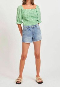 Zielony top z bufiastymi rękawami i marszczonym wzorem, zestawiony z jasnoniebieskimi szortami dżinsowymi. Model nosi beżowe, puszyste sandały.