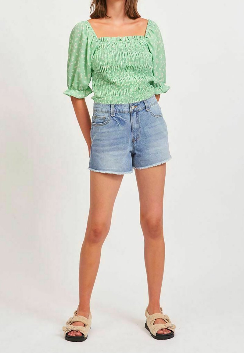 Zielony top z bufiastymi rękawami i marszczonym wzorem, zestawiony z jasnoniebieskimi szortami dżinsowymi. Model nosi beżowe, puszyste sandały.