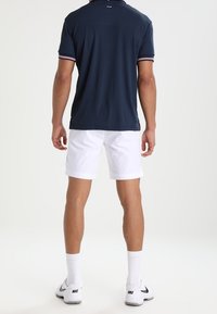 Muž v námořnickém polu, bílých šortkách a bílých teniskách Nike, stojí otočený zády proti jednoduchému bílému pozadí.