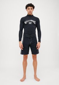 Czarna, długoznachowa koszulka do pływania z białym logo "Billabong", w połączeniu z czarnymi szortami do boardingu z białymi szwami i wiązanym paskiem w talii.