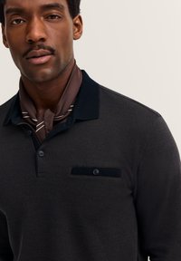 Dunkel gemustertes Langarm-Polo mit marineblauem Kragen, Zweiknopf-Leiste und einer aufgenähten Tasche mit Knopfdetail. Brauner Schal mit Streifen.