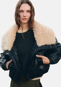 Giacca bomber in pelle nera con colletto in pelliccia sintetica beige, chiusura con zip e polsini a costine. Indossata sopra un maglione scuro.