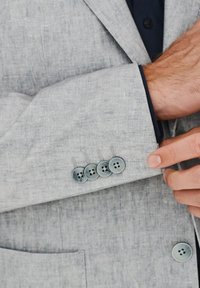 Jasnoszary żakiet z teksturowanej tkaniny, z czterema niebieskimi guzikami na mankiecie i niebieską podszewką widoczną w otworze rękawa.