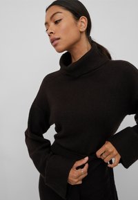 Pullover de lã castanho escuro com colarinho alto, mangas largas e cintura cingida. Textura suave com um ajuste descontraído.