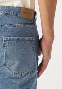 Jean en denim bleu clair avec un patch en cuir portant l'inscription "SAMSE." Comprend une poche arrière avec une couture marquée et un tissu texturé.
