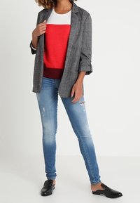Blazer gris de cuadros sobre un suéter bloqueado en rojo y blanco, combinado con jeans claros desgastados y zapatos negros tipo slip-on.