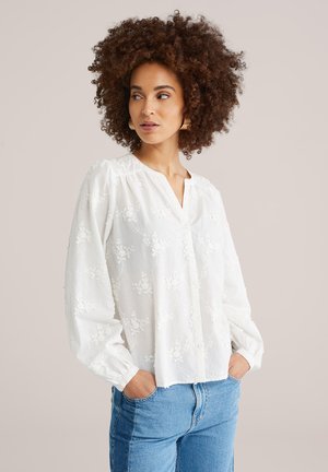 Frau mit lockigem Haar, die goldene Creolen, eine weiße, langärmelige Bluse mit Blumenstickerei und blaue Jeans trägt, mit den Händen in den Taschen.