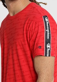 Rød T-shirt med korte ærmer og gentagne "Champion" logo mønster, med en sort og hvid logo stribe langs skulderen.