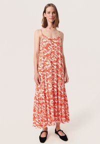 Maxikleid in einem fließenden Design, mit einem orange-weißen abstrakten Muster, dünnen Trägern und einem gestuften Rock. Trägt es mit schwarzen Ballerinas.