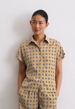 Chemise crème à manches courtes, ornée d'un motif géométrique bleu et noir. Design à col avec patte de boutonnage et coupe décontractée.