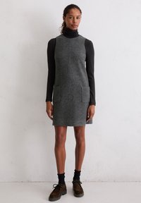 Grijze wollen mouwloze jurk met hoge halslijn, twee voorzakken en een rechte snit, gecombineerd met een zwart longsleeve shirt en donkere schoenen.