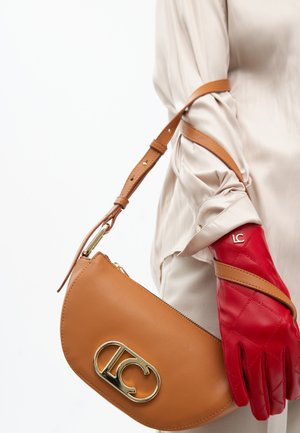 Eine Person trägt eine beige Bluse und hält eine braune Handtasche mit einem goldenen Logo, während sie einen roten Lederhandschuh trägt. Der Hintergrund ist weiß.