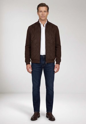 Mann steht und trägt eine braune gesteppte Jacke, weißes Hemd, dunkelblaue Jeans und braune Lederstiefel vor einfachem Hintergrund.