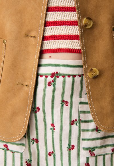 Giacca in ecopelle color beige con due bottoni, indossata sopra un top a righe rosso e crema in maglia, abbinata a una gonna in denim con ricami floreali e righe.