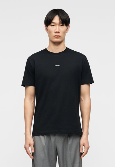 Νεαρός άνδρας με ίσια μαύρα μαλλιά φοράει εφαρμοστό μαύρο μπλουζάκι με το κείμενο "ICEBERG" και γκρι πιετέ παντελόνια σε απλό φόντο.