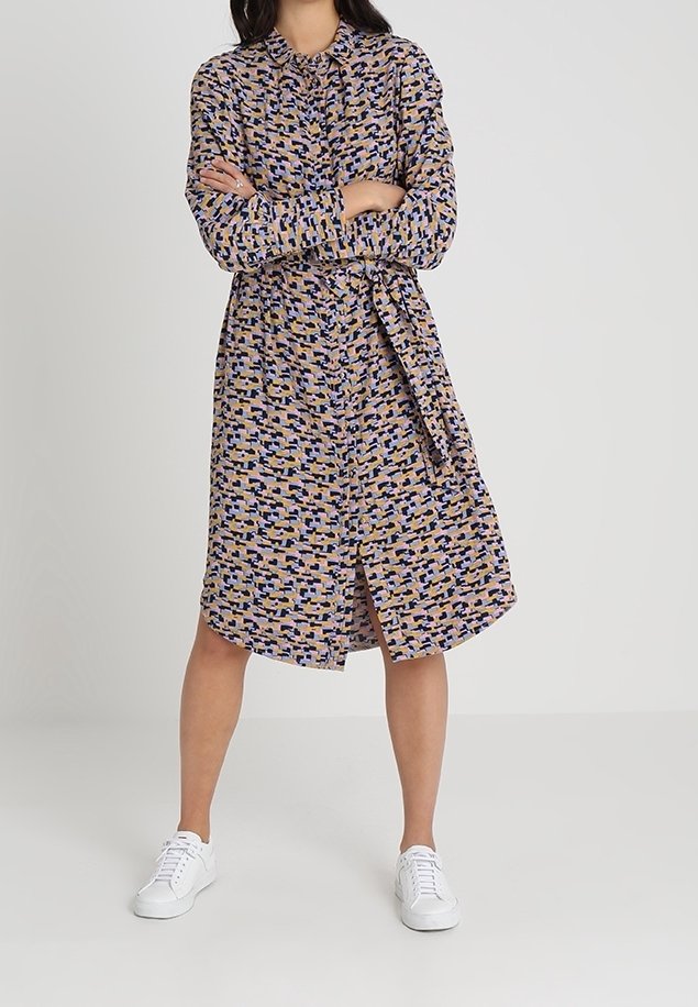 Femme portant une robe-chemise à motifs, manches longues, ceinturée, et des baskets blanches, debout les bras croisés sur un fond uni.
