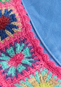 Bestickter Stoff mit mehrfarbigen Blumenmustern in Pink, Blau, Grün und Lila, vor einem glatten, hellblauen Hintergrund.
