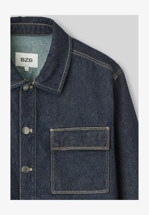 Veste en denim bleu foncé avec un col, fermeture à boutons et une poche à patch. Présente des coutures contrastées et un tissu texturé.