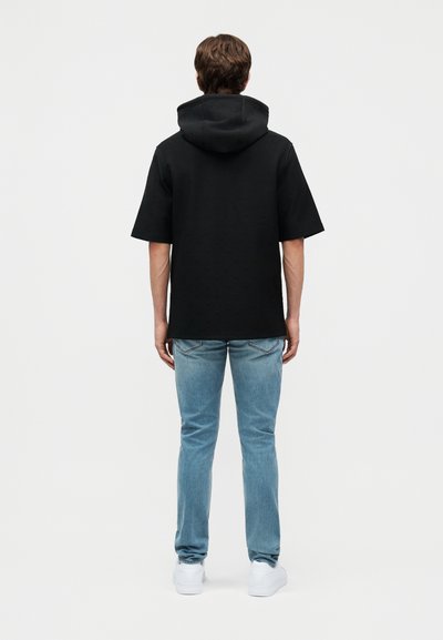 Άνδρας που στέκεται με την πλάτη γυρισμένη, φοράει μαύρη κοντομάνικη μπλούζα με κουκούλα, ανοιχτό μπλε τζιν και λευκά αθλητικά σε απλό λευκό φόντο.
