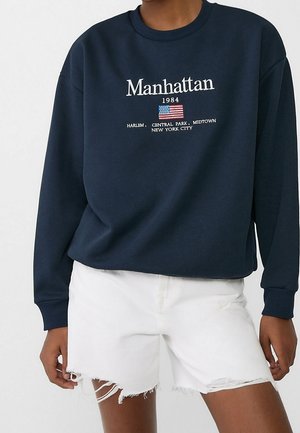 Person trägt einen marineblauen Pullover mit der Aufschrift "Manhattan 1984" und einer kleinen amerikanischen Flagge, kombiniert mit weißen, ausgefransten Denim-Shorts.