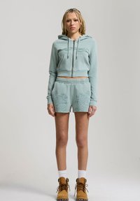 Jeune personne aux cheveux blonds tressés portant un sweat à capuche bleu clair zippé, un short assorti brodé de fleurs, des chaussettes blanches et des bottes beige.