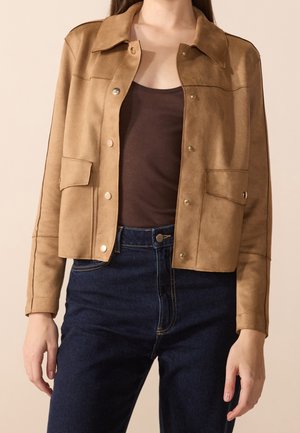 Femme portant une veste beige boutonnée sur un débardeur marron, associée à un jean bleu foncé taille haute, sur un fond uni.