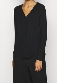Top lungo nero con maniche a lungo e scollatura a V, realizzato in tessuto liscio. Presenta un design plissettato sul davanti e un orlo curvo.