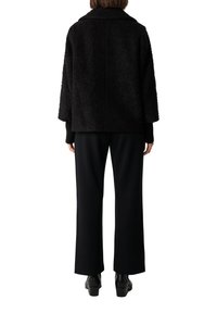 Giacca nera con texture, collo ampio, vestibilità over, spalle scese, abbinata a pantaloni neri a gamba larga e stivali alla caviglia.