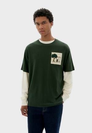 Green cotton t-shirt layered over a white long-sleeve top, featuring a pocket with embroidered tree and architectural details.
