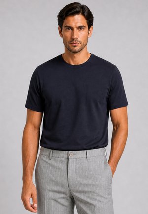 Mann mit dunklen Haaren, der ein schlichtes schwarzes Kurzarm-T-Shirt und graue Hose mit Nadelstreifen trägt, steht mit einer Hand in der Tasche.