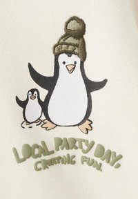 Tissu crème avec un motif brodé représentant deux pingouins. Le pingouin le plus grand porte un chapeau vert ; le texte indique "JOUR DE FÊTE LOCALE, CRÉER DU PLAISIR."