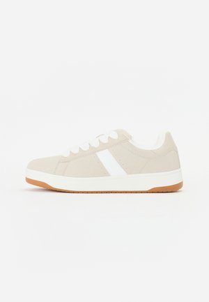 Beige sneaker with white accents, suede texture, rounded shape, lace-up design, and a contrasting gum rubber outsole.