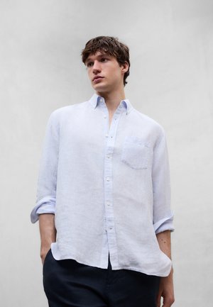 Joven con camisa de rayas azul claro de manga larga con las mangas remangadas, de pie con las manos en los bolsillos de pantalones oscuros frente a un fondo liso.