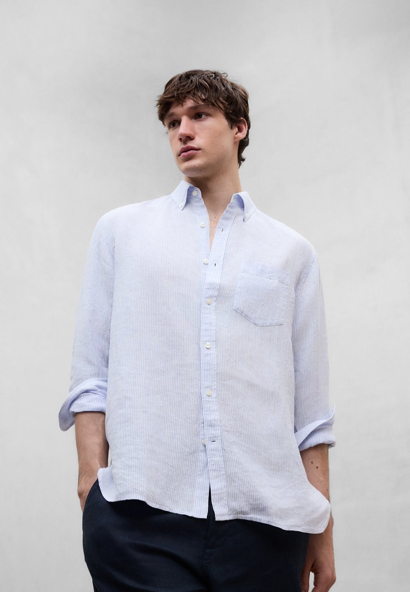 Jeune homme portant une chemise à manches longues rayée bleu clair avec les manches retroussées, debout les mains dans les poches d'un pantalon foncé, devant un fond uni.