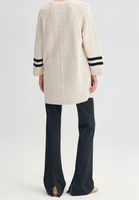 Persona che indossa un pullover a maglia beige texture con strisce nere sulle maniche e pantaloni larghi neri con spacco sul retro sopra scarpe beige.
