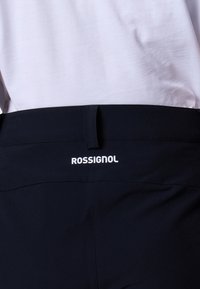 Gros plan sur une personne portant un pantalon noir avec le logo Rossignol sur la ceinture et une chemise blanche rentrée.