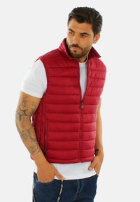 Gilet rosso gonfio con colletto alto e chiusura con zip. Presenta una texture trapuntata e due tasche laterali. Indossato sopra una maglietta bianca.