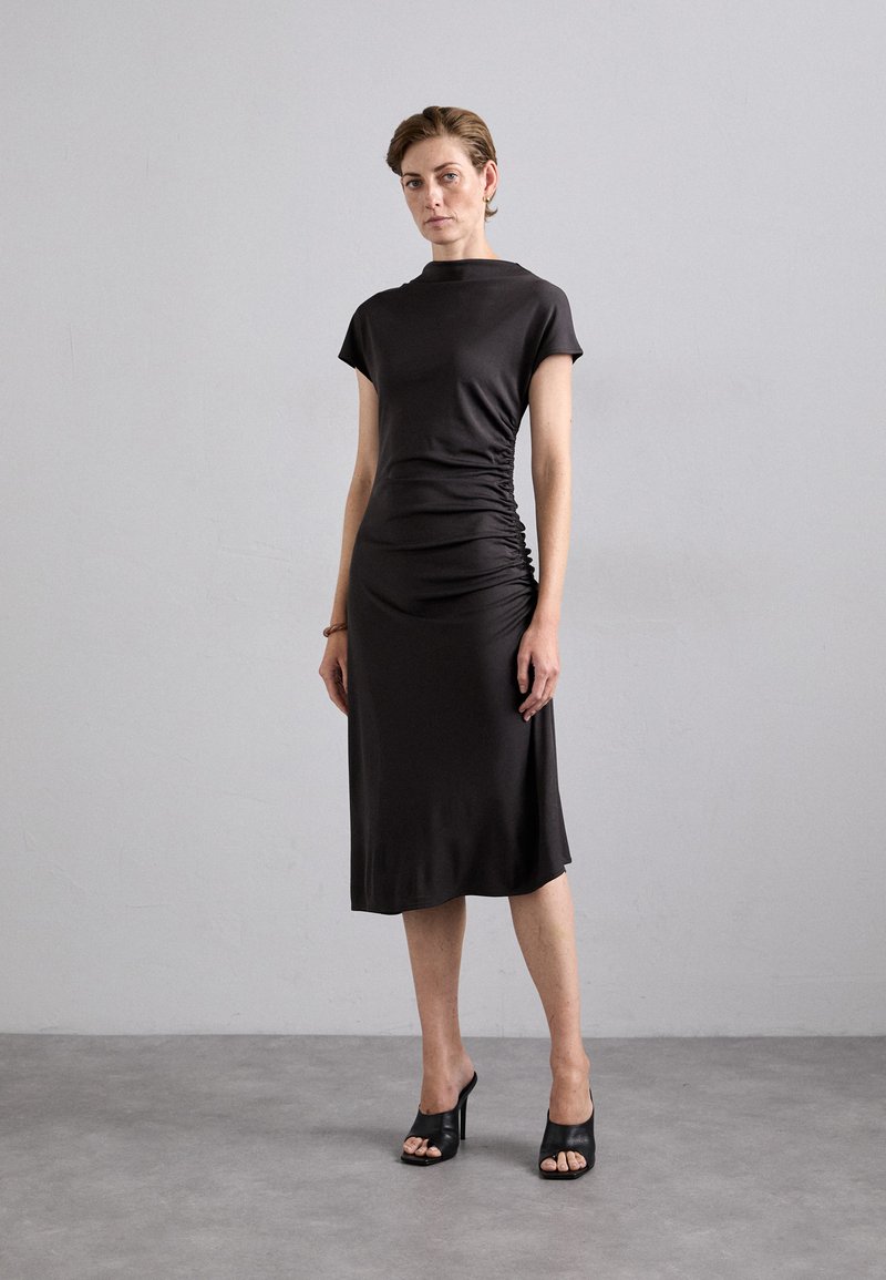 Fitted, knee-length dress in dark gray fabric with short sleeves and side ruching details. Worn with open-toe black heels.
